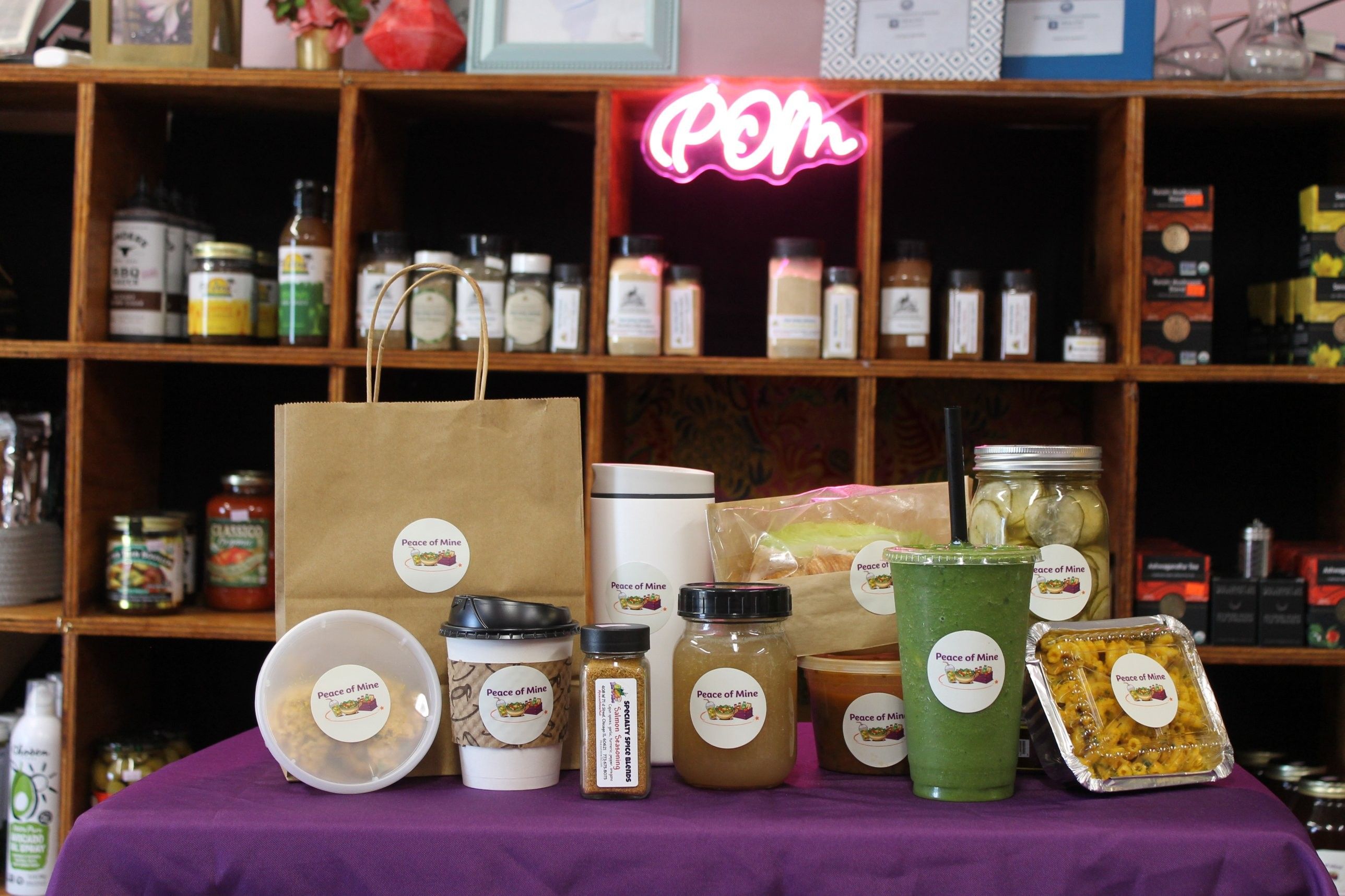 Peace of Mine soup, coffee, spice blend, smoothie, pickles and mac and cheese on a purple table under the pink neon POM sign